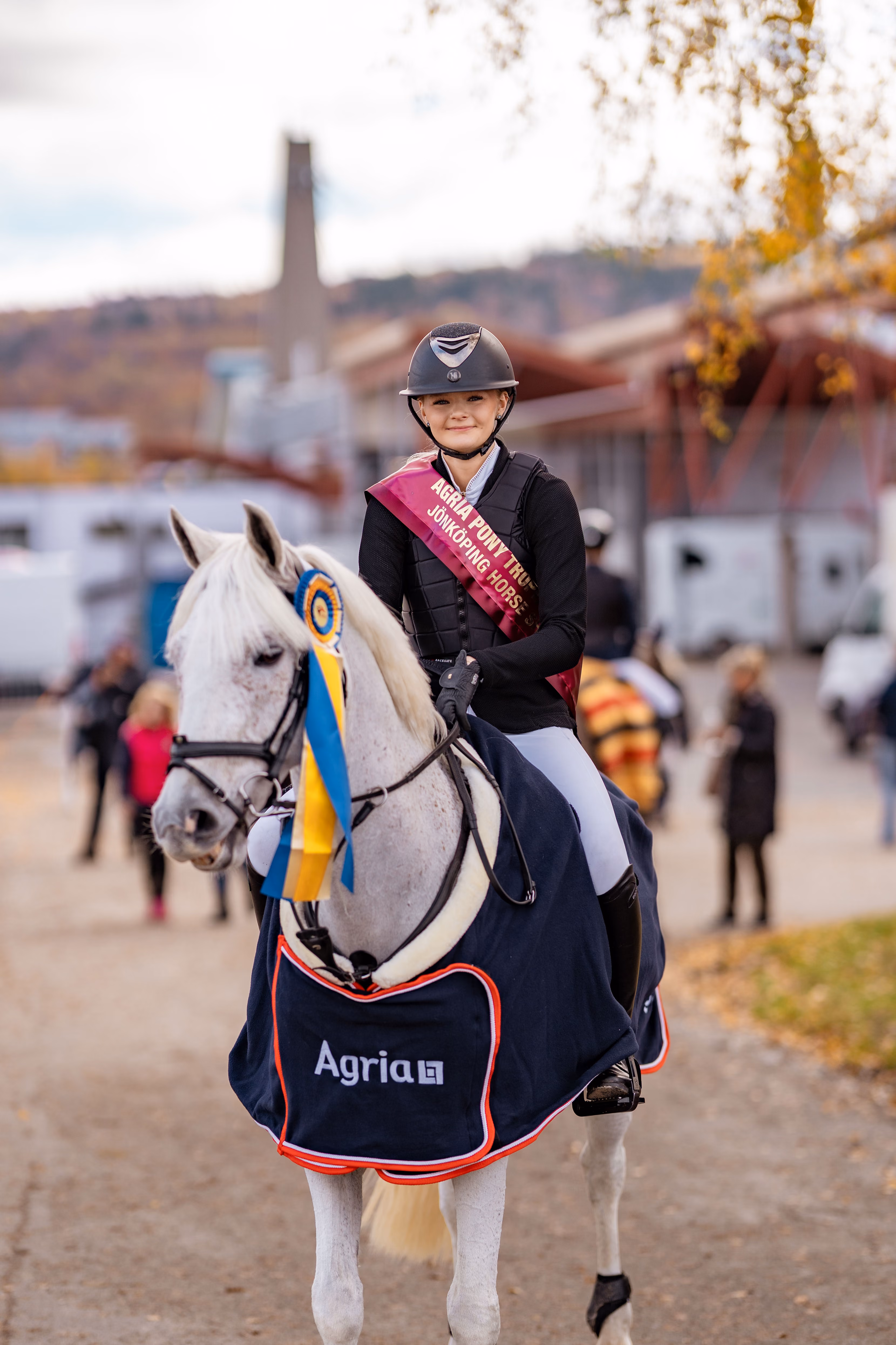 Smiling rider wearing a trophy sash on a white horse with ribbons and an Agria blanket.