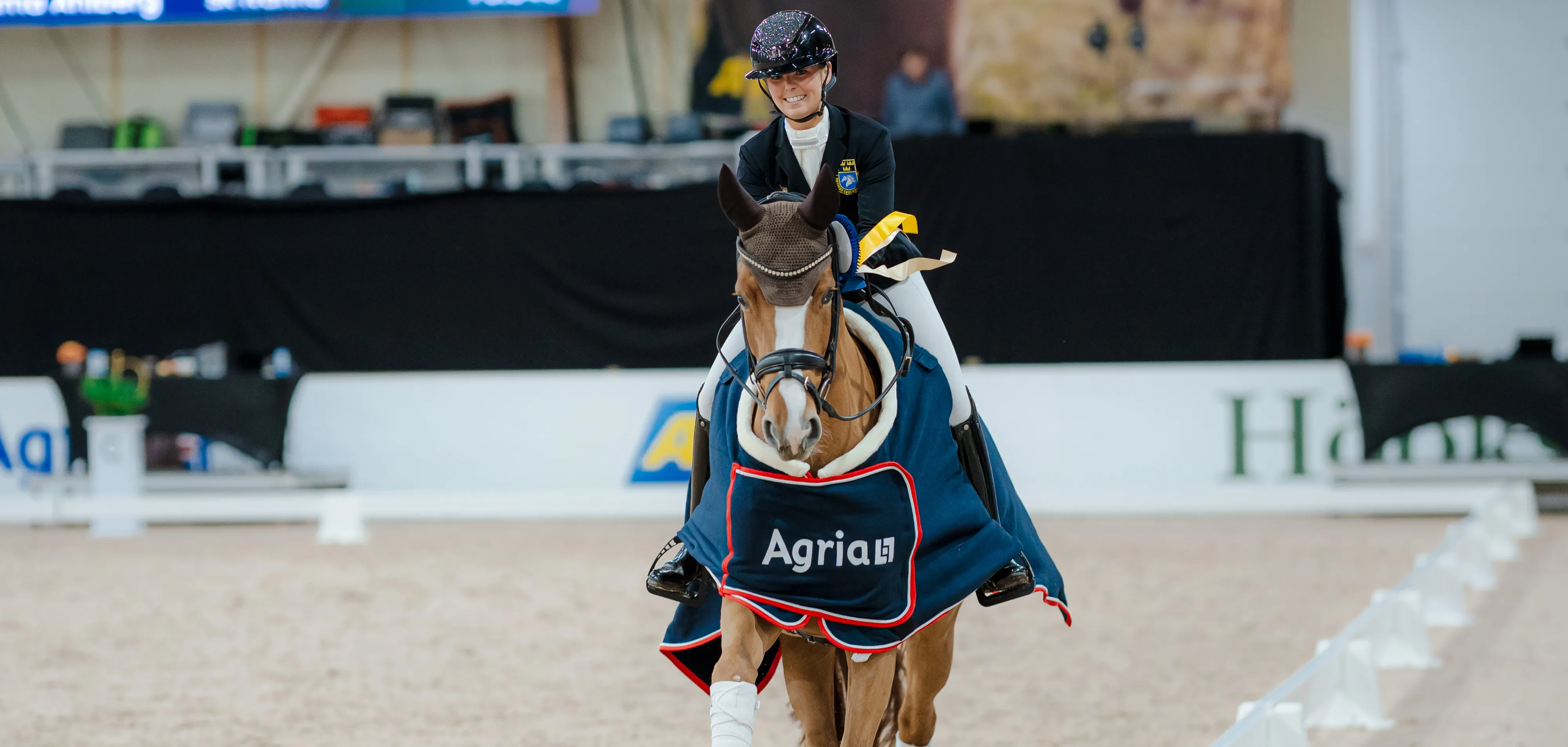 Smiling equestrian rider in formal gear on a horse with Agria blanket and ribbon in arena.