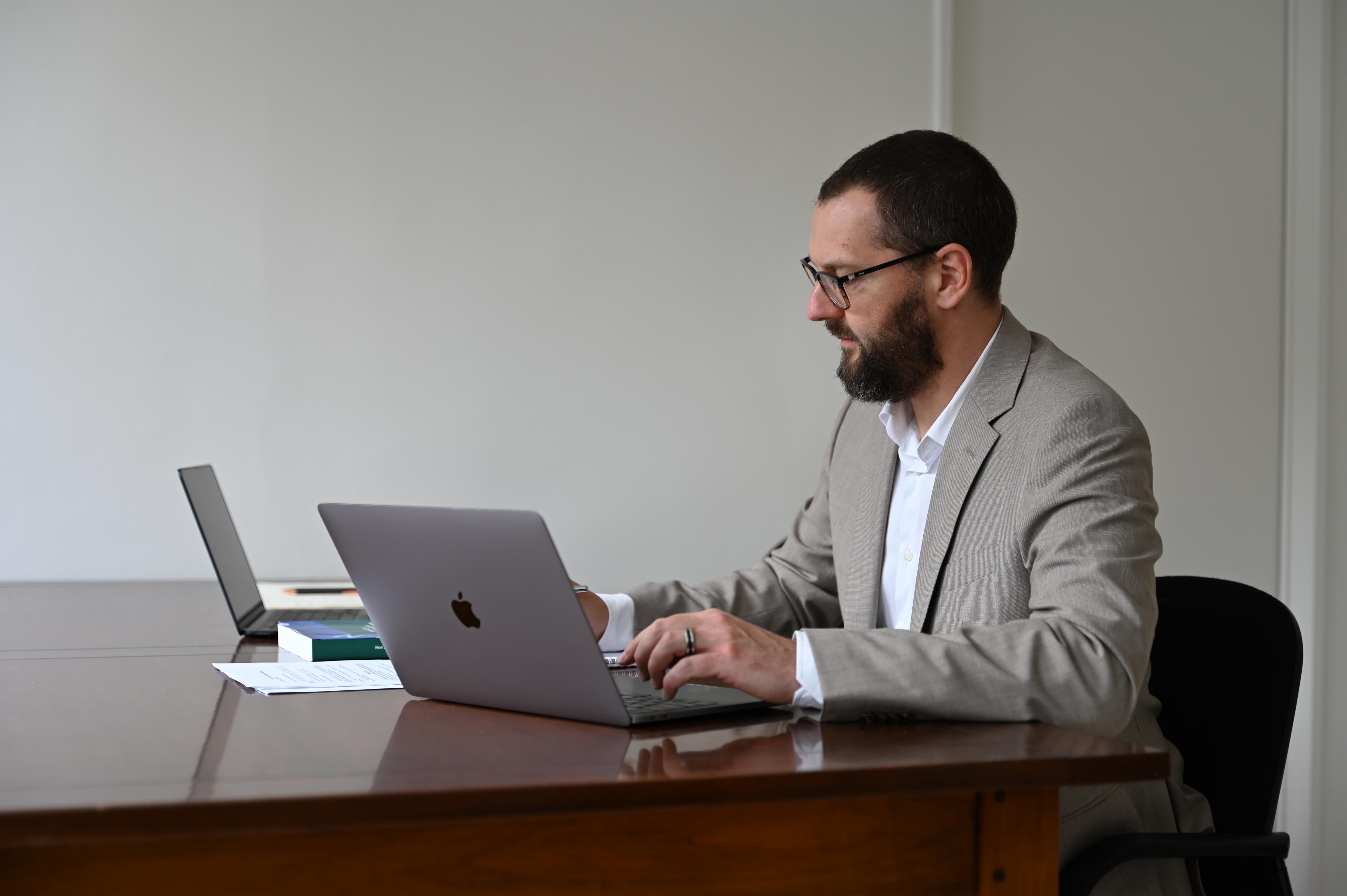 Man met bril en grijs pak werkt op een laptop aan een houten tafel met papieren en een tweede laptop.