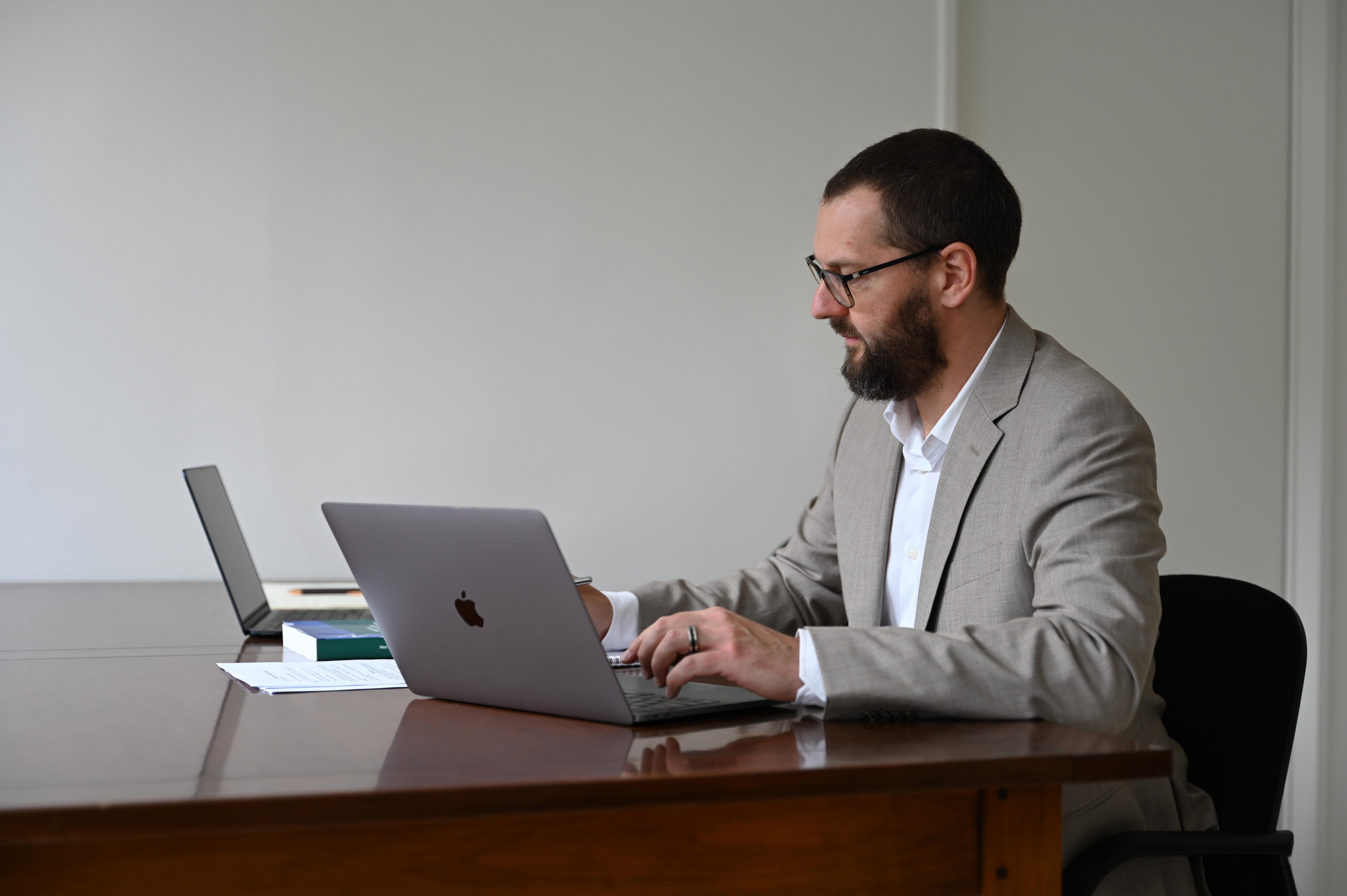 Man in beige suit and glasses werkt zittend aan een bureau op een laptop met documenten en boeken.