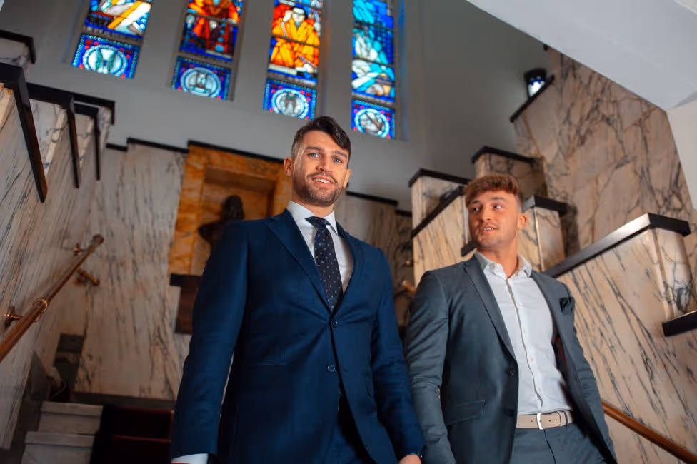 Twee mannen in pakken lopen een trap op met marmeren wanden en glas-in-loodramen.