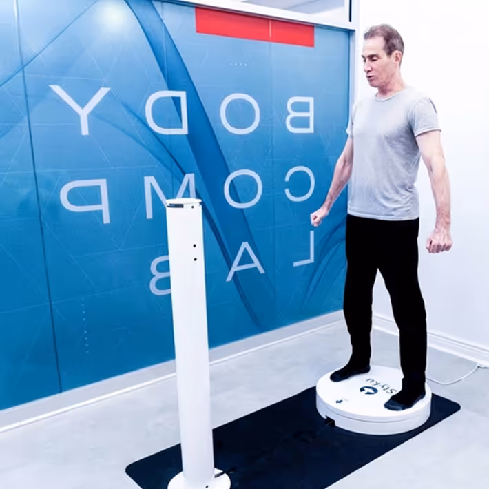 Man standing on a white circular vibration exercise platform in a lab with 'BODY COMP LAB' text on the blue wall behind him.