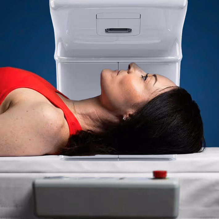 Woman lying on her back wearing a red tank top, positioned for a medical scan with a white scanning machine above her head.