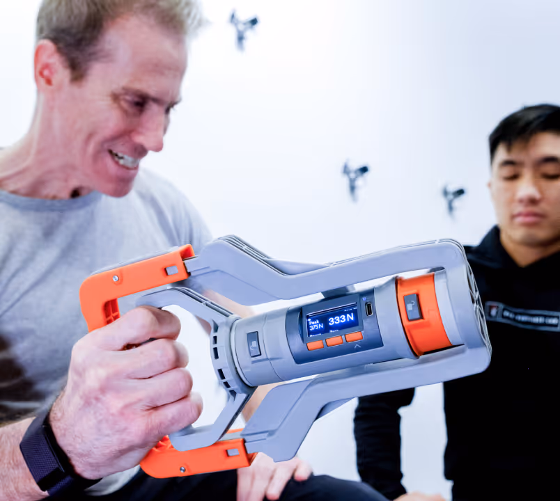 Man holding a gray and orange digital grip strength measuring device displaying 333N, with another man observing in the background.