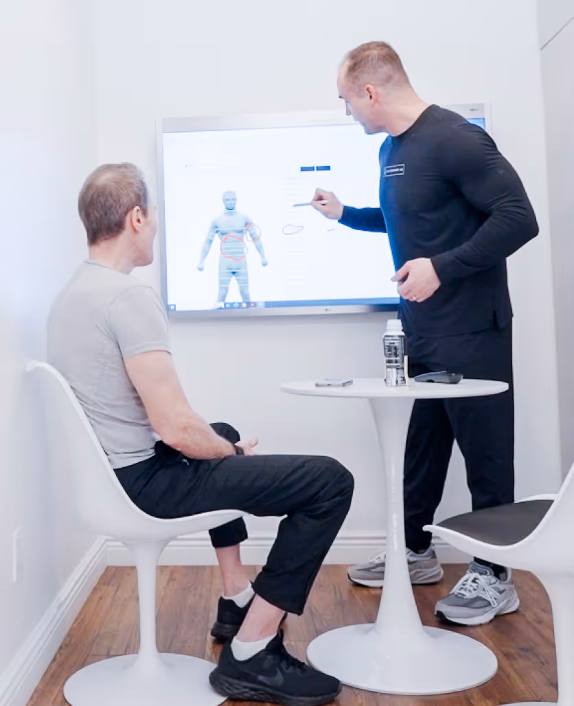 Two men in a small room with one seated and the other standing, discussing a digital human anatomy model displayed on a wall-mounted screen.