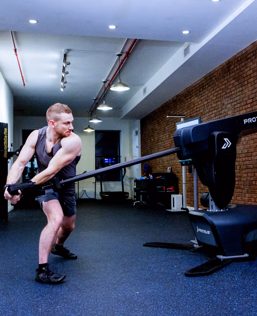 Man with muscular arms using a resistance training machine in a gym with brick wall and ceiling lights.