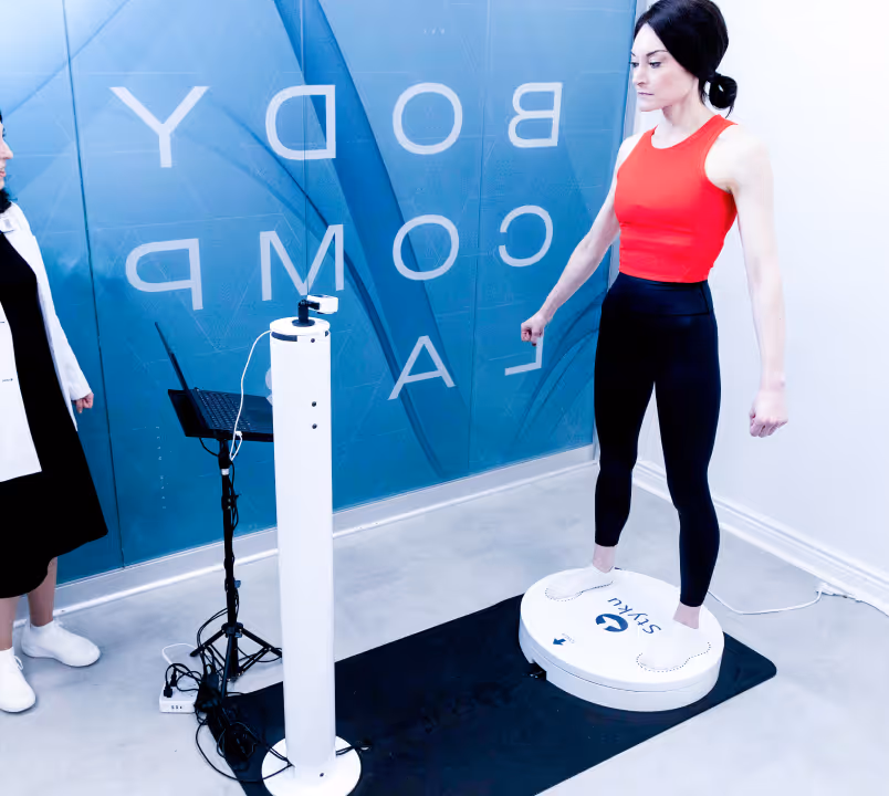 Woman in red tank top and black leggings standing on a white foot-shaped body composition scanner while a person in a white coat stands nearby.