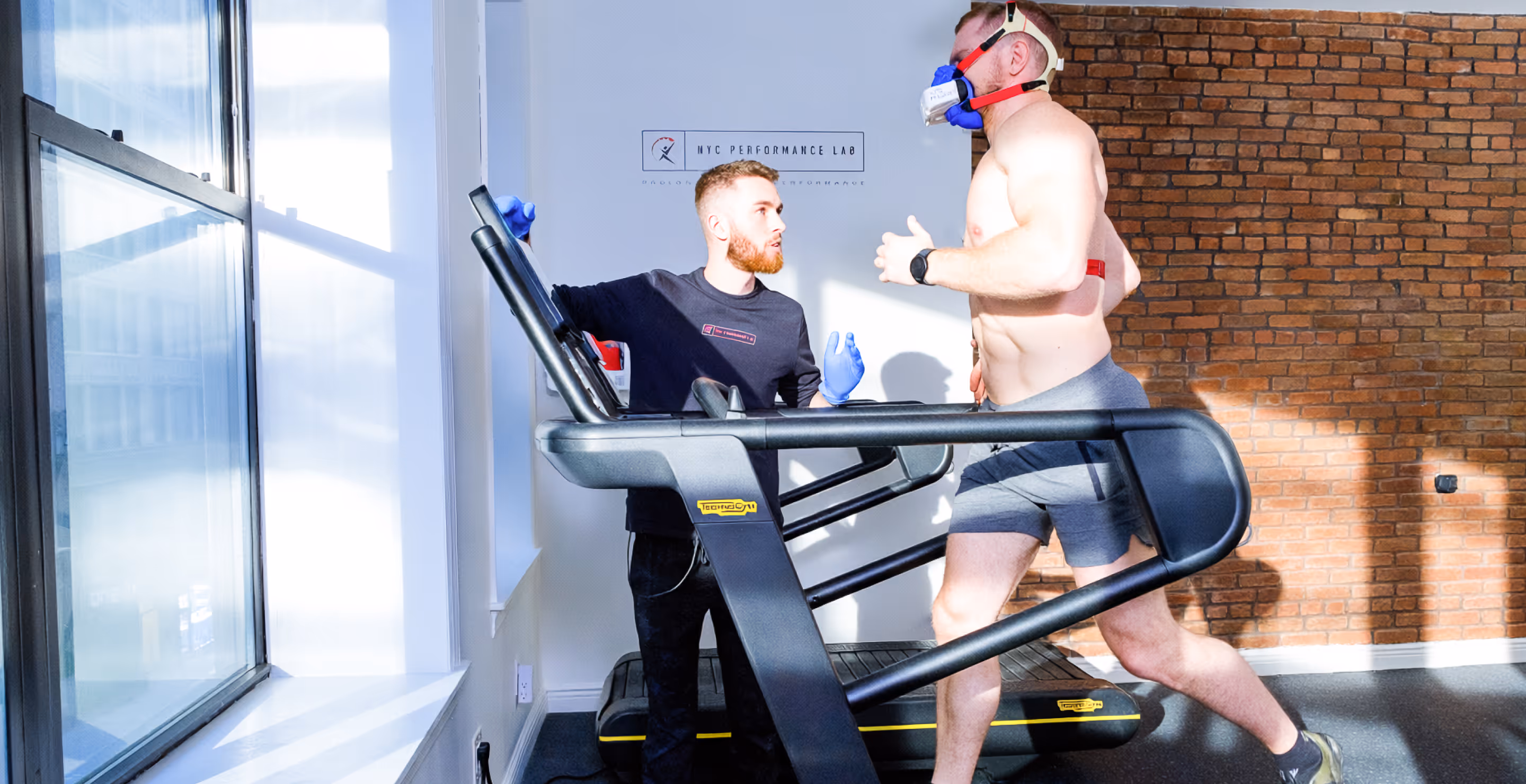 Man running on treadmill wearing a respiratory mask while a trainer in gloves monitors and instructs him inside a gym with a brick wall background.