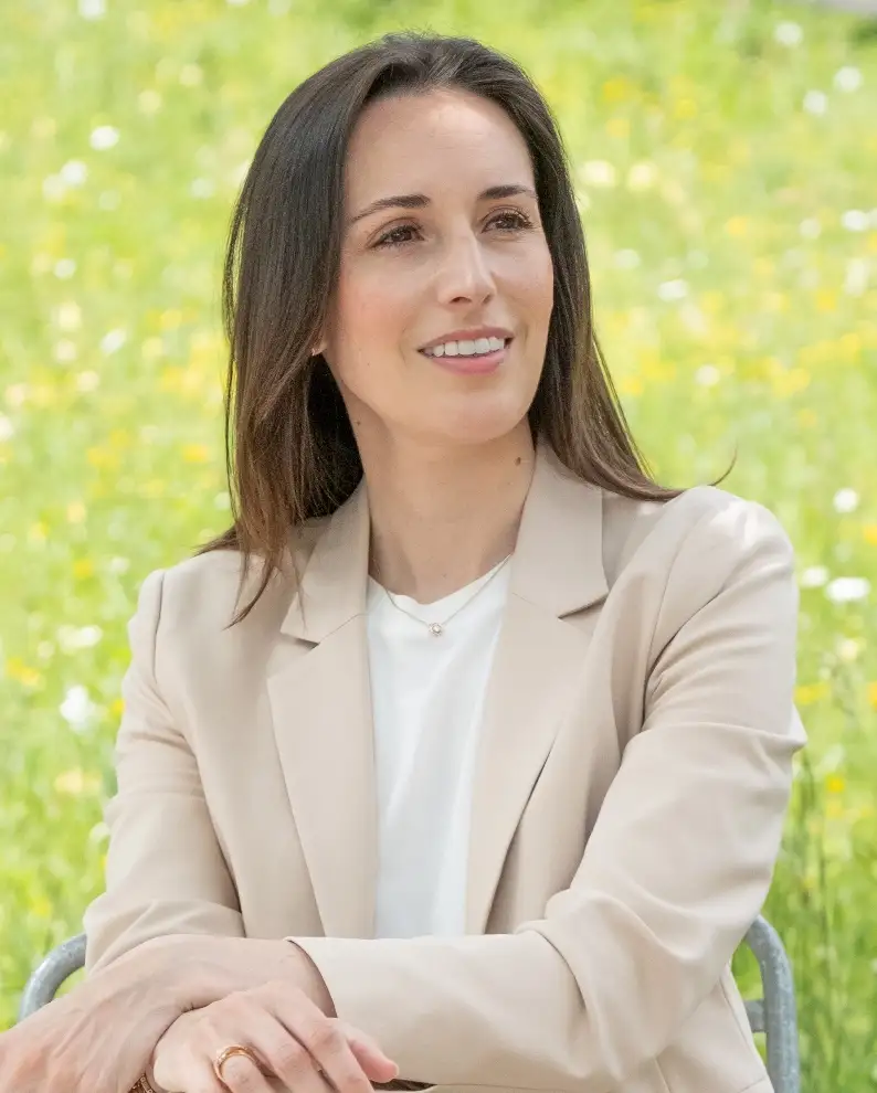 Karin Stadelmann trägt einen hellbeigen Blazer und sitzt entspannt vor grüner Wiese mit Blumen.