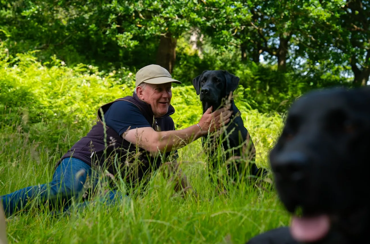 A man crouches in long grass with a black Labrador retriever, gently holding the dog's collar as they share a moment together outdoors.