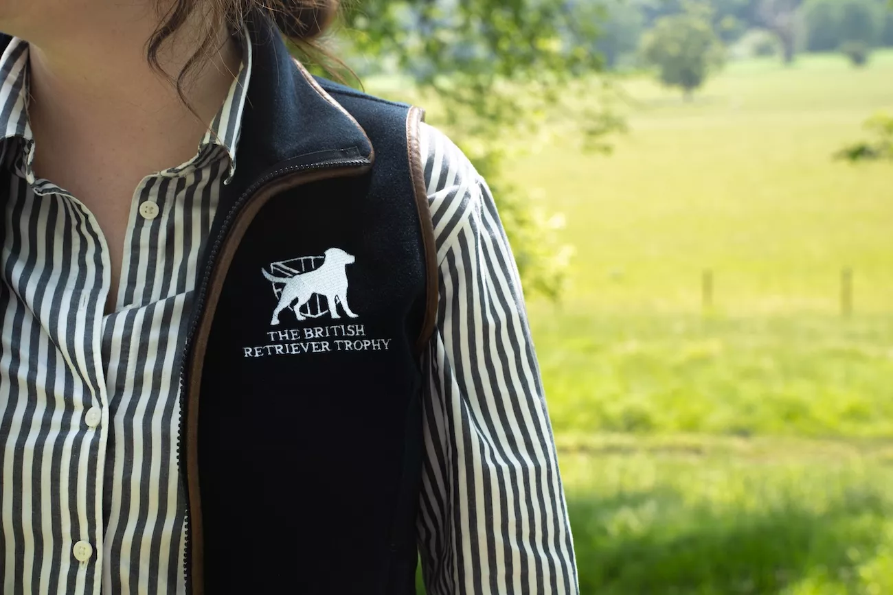 Close-up of The British Retriever Trophy embroidered logo on a navy fleece gilet, worn over a striped shirt with countryside fields in the background.