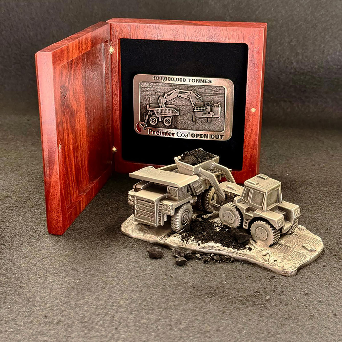 Custom mining recognition display featuring a pewter front end loader and haul truck scene with coal detail, presented alongside a Premier Coal Open Cut 100,000,000 tonnes commemorative buckle in a handcrafted timber display box.