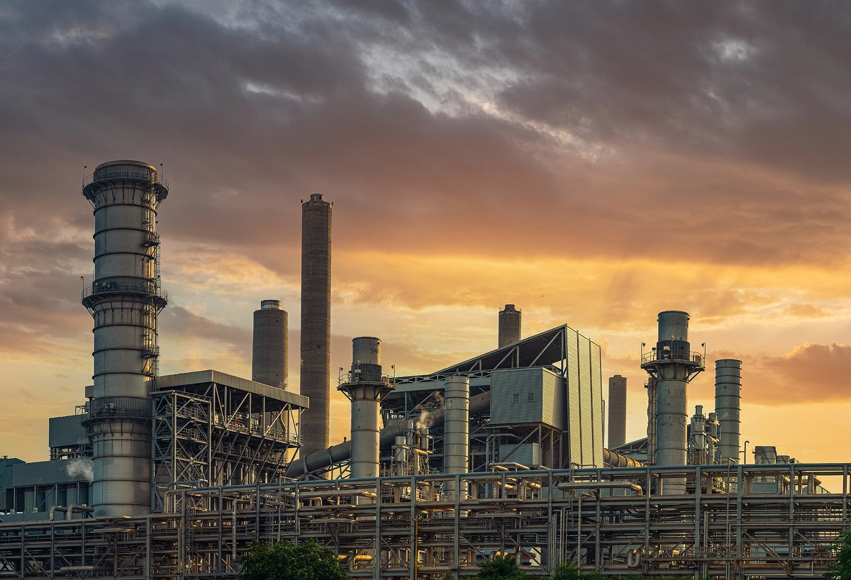 Industrial factory with multiple smokestacks and complex piping under a dramatic sunset sky.