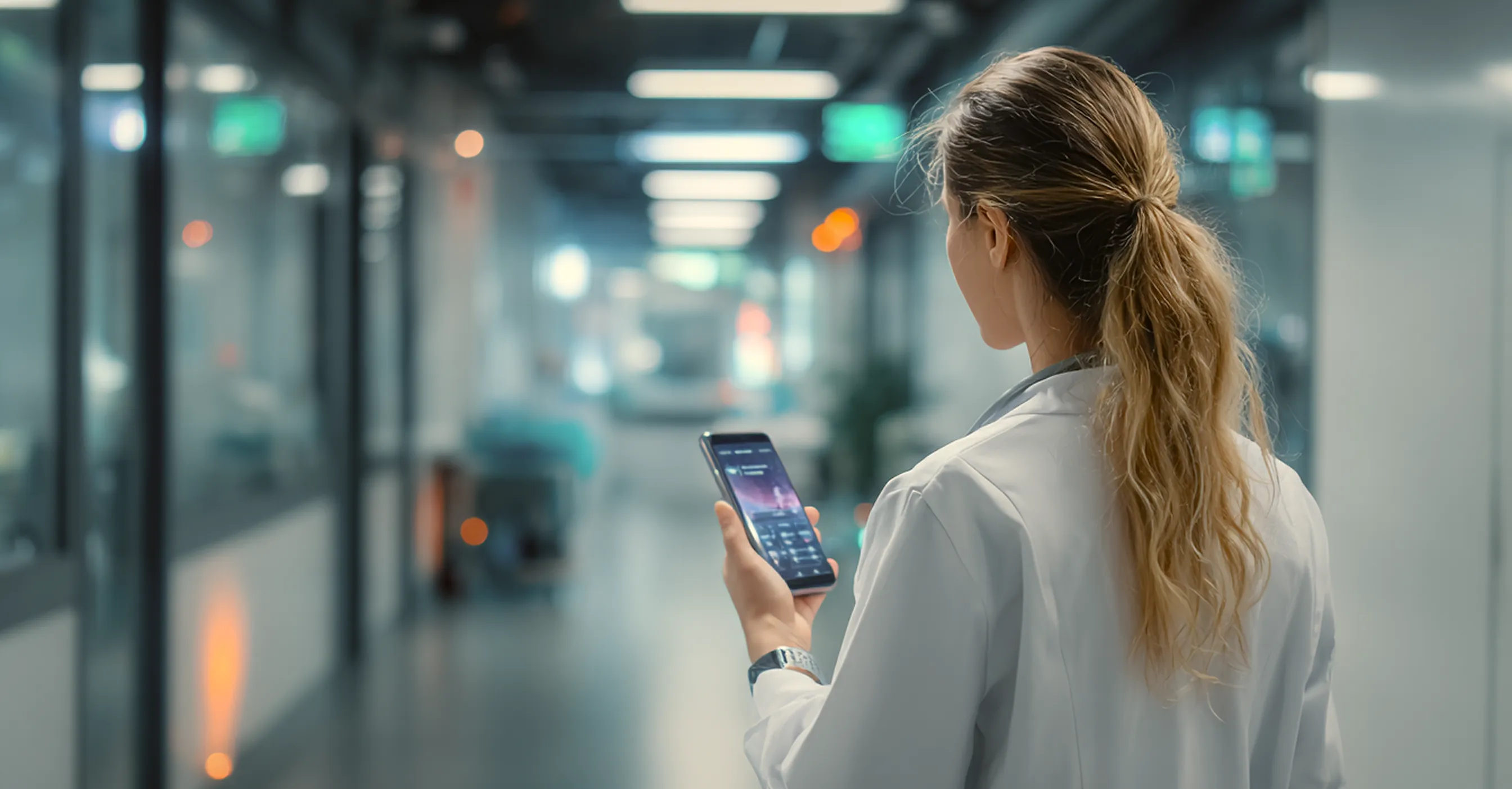Medical professional in white coat looking at a smartphone in a modern hospital corridor.