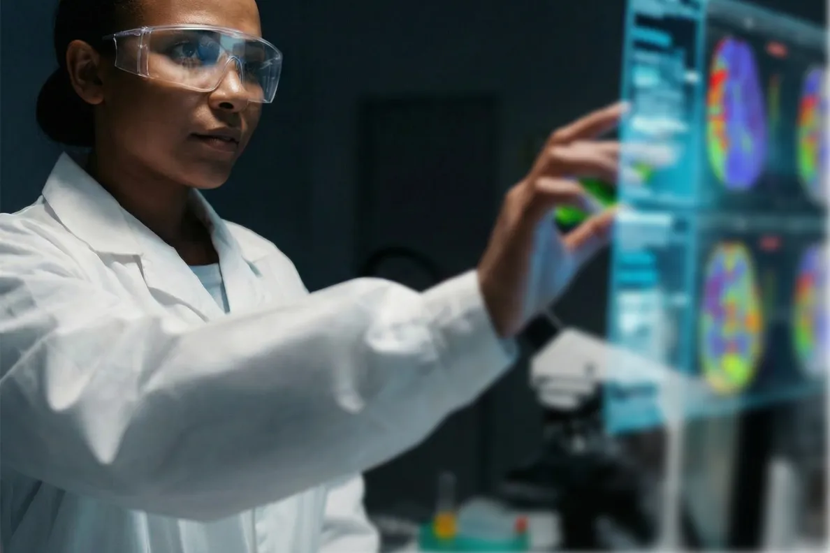 Scientist wearing safety glasses uses touchscreen displaying colorful brain scan images.
