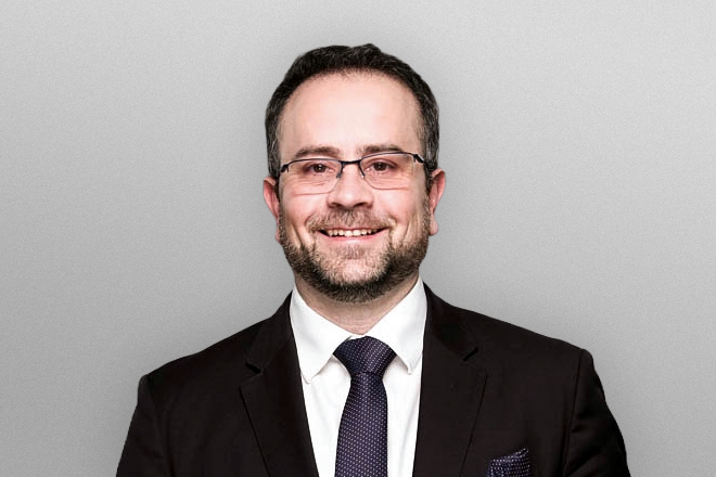 Smiling man with glasses in a black suit, white shirt, and dark tie against a gray background.
