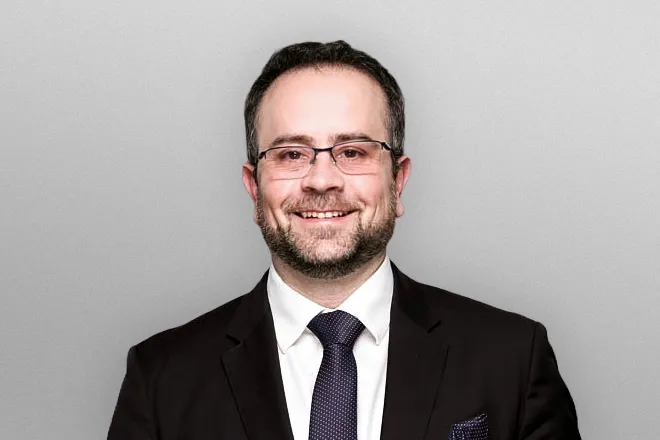 Smiling man with glasses in a black suit, white shirt, and dark tie against a gray background.