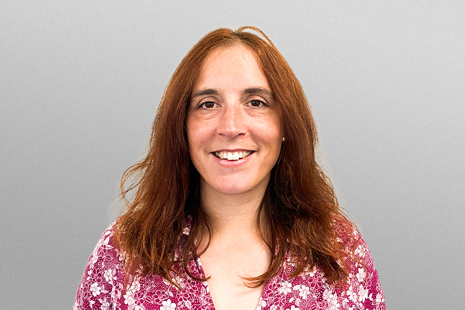 Smiling woman with long reddish hair wearing a pink floral shirt against a gray background.