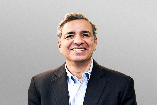 Smiling man with graying hair wearing a dark blazer and light blue shirt against a gray background.