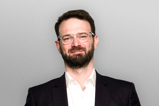 Man with glasses and beard wearing a white shirt and black suit jacket against a gray background.