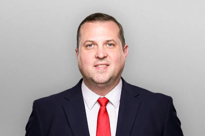 Man in dark suit, white shirt, and red tie smiling against a plain gray background.