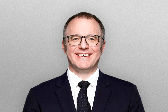 Smiling man wearing glasses, black suit, white shirt, and dark tie against a gray background.