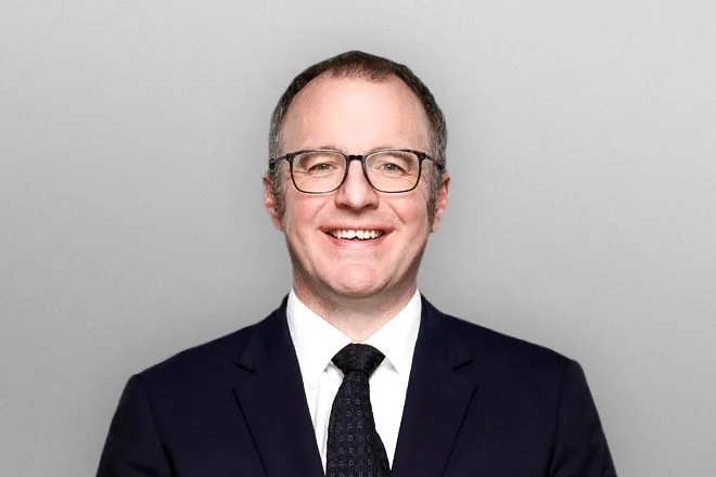 Smiling man wearing glasses, black suit, white shirt, and dark tie against a gray background.