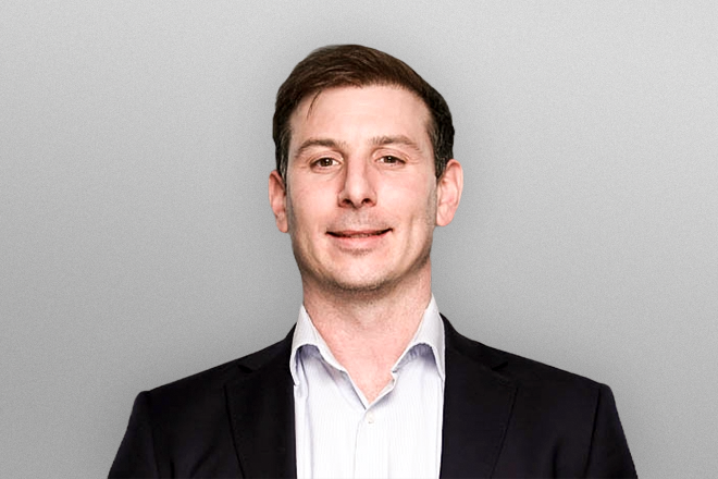 Man with short brown hair wearing a white shirt and black blazer against a gray background.