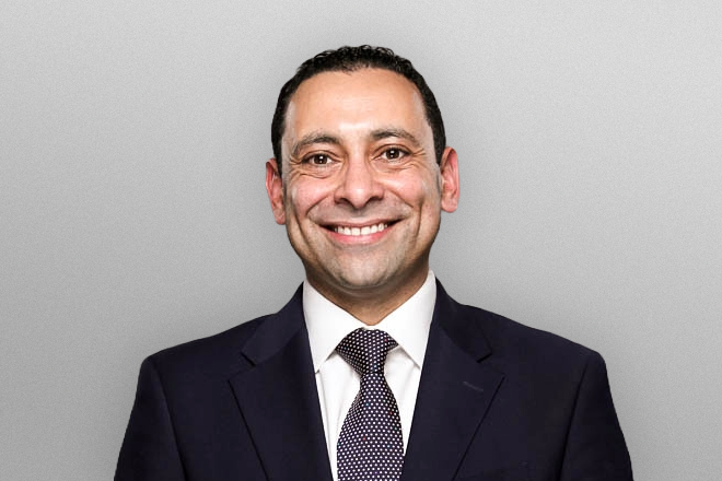 Smiling man in a dark suit, white shirt, and patterned tie against a plain light gray background.