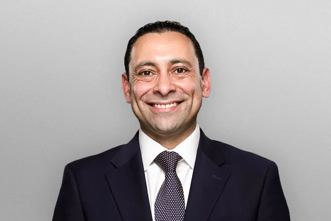 Smiling man in a dark suit, white shirt, and patterned tie against a plain light gray background.