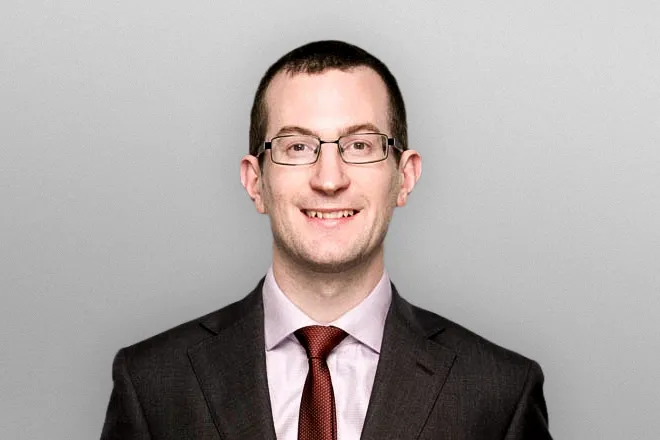 Smiling man wearing glasses, a dark suit, light shirt, and red tie, against a plain gray background.