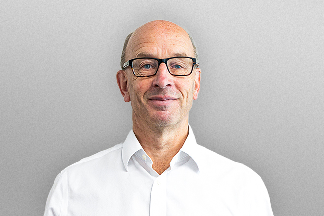 Middle-aged man with glasses wearing a white shirt standing against a gray background.