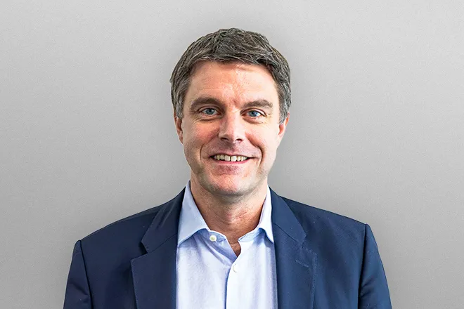 Smiling man with blue eyes wearing a navy blazer and light blue shirt against a gray background.