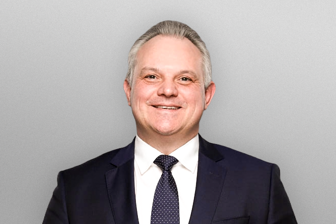 Smiling middle-aged man in dark suit and polka dot tie against a light gray background.