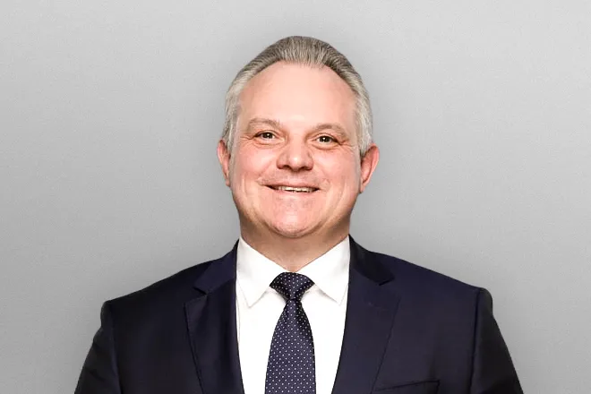 Smiling middle-aged man in dark suit and polka dot tie against a light gray background.