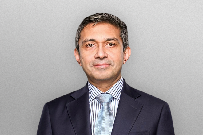 Middle-aged man with short dark hair wearing a dark suit, striped shirt, and light blue tie on gray background.