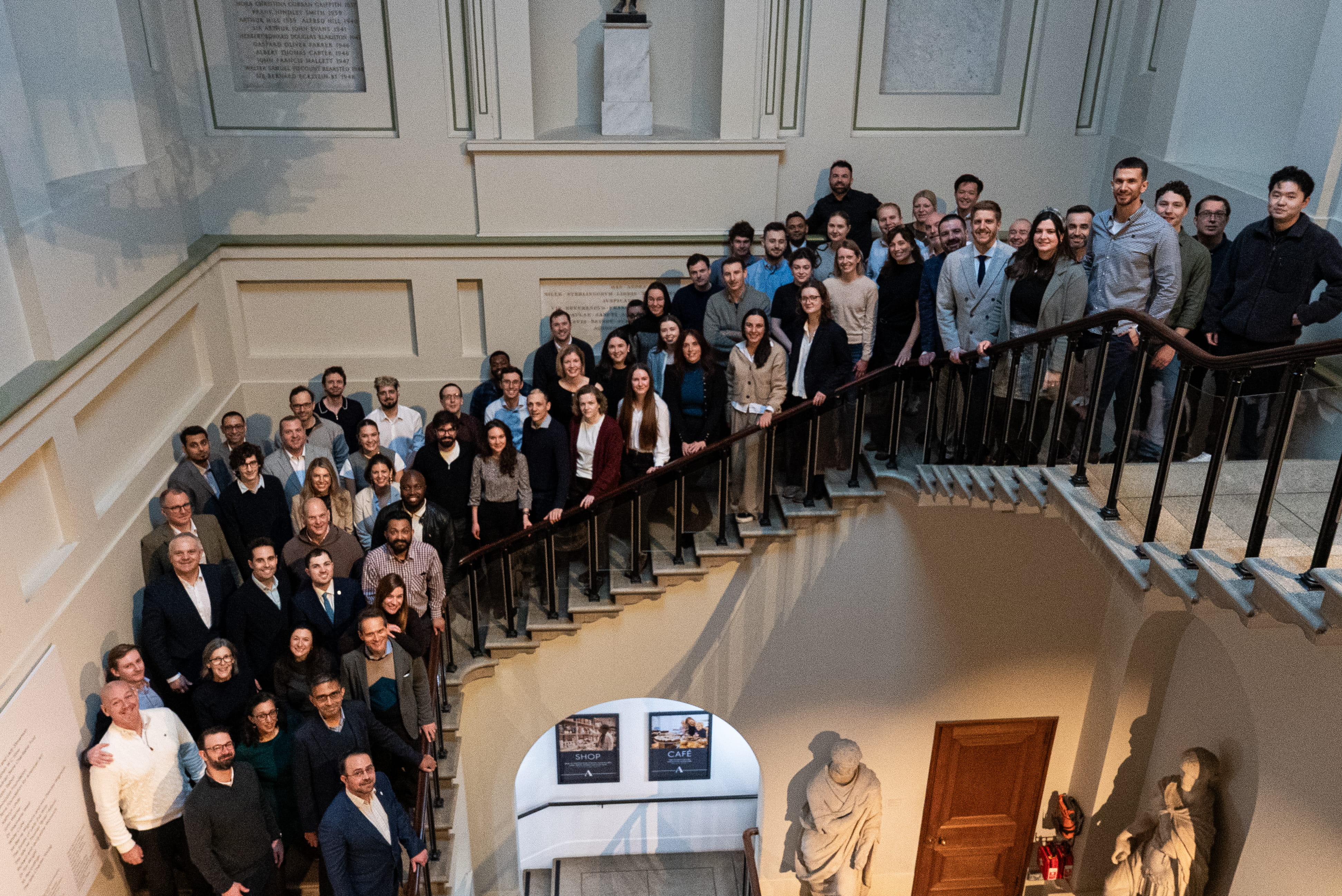 Large group of people posing on a staircase inside a building with classical statues below.