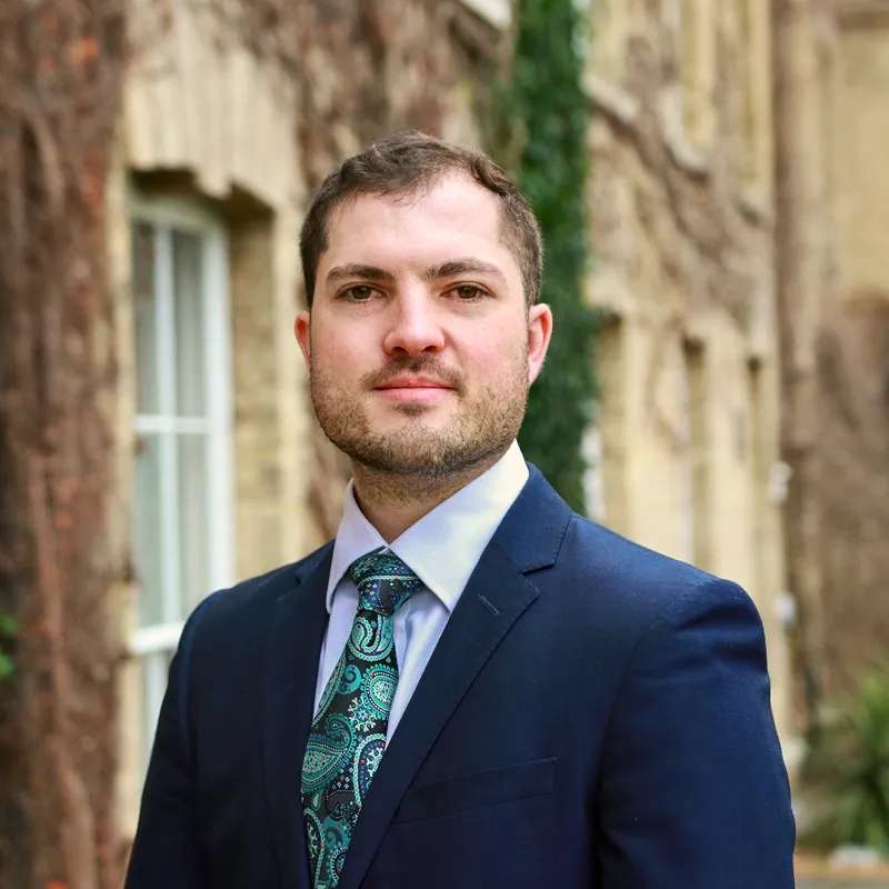 Man in blue suit with paisley tie standing outside near stone building with window.