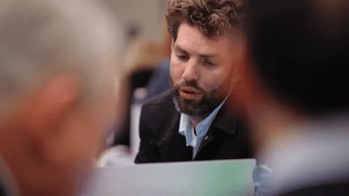 Bearded man with curly hair speaking at a meeting while others listen from blurred foreground.