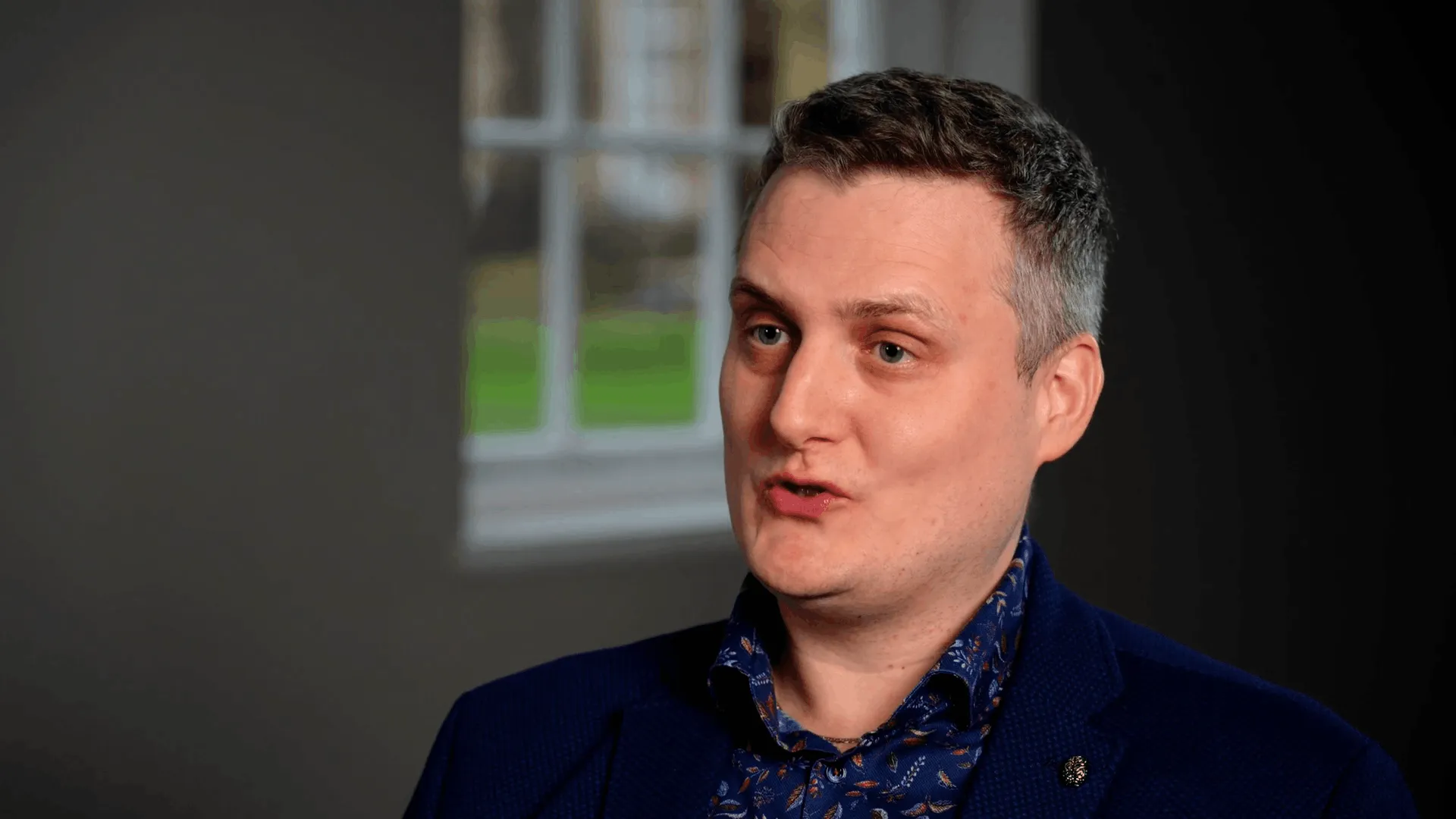 Man with short hair speaking, wearing a blue floral shirt and navy blazer, blurred window background.