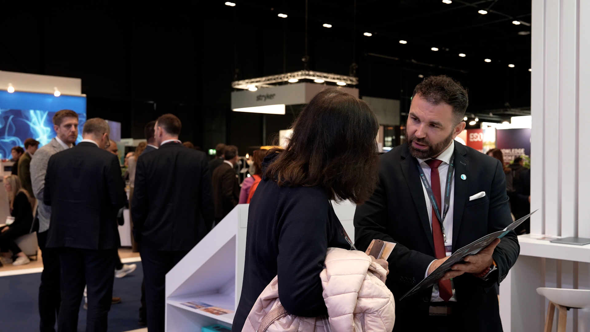 Man in suit showing a brochure to a woman holding a pink jacket at a busy exhibition booth.