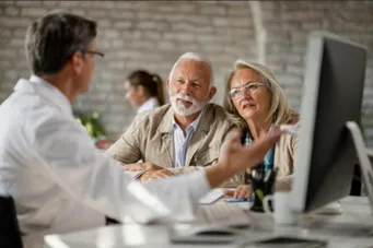 Doctor discussing medical information on a computer with an elderly couple in a bright office.