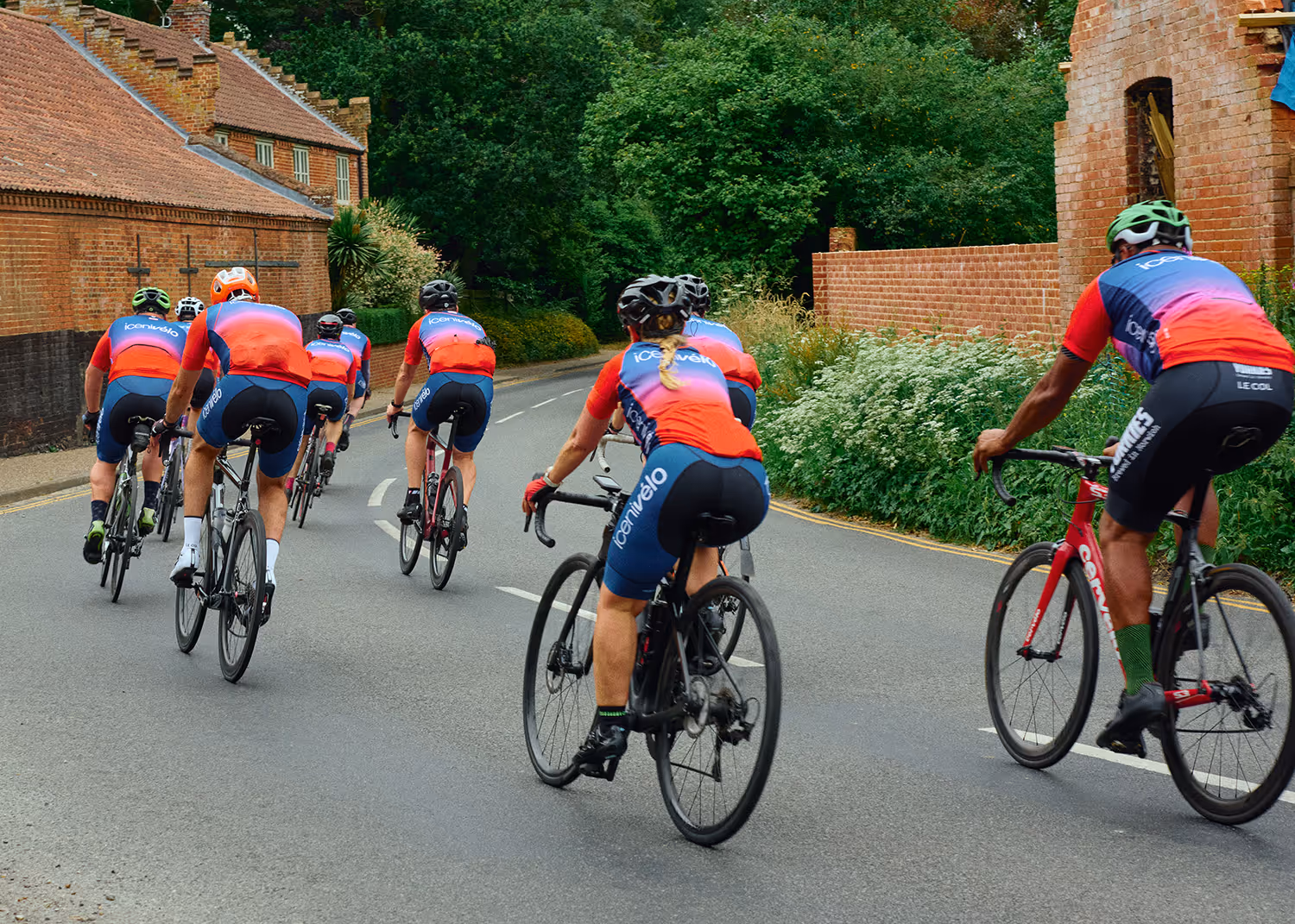 Group of cyclists in matching red and blue jerseys riding on a road past brick buildings and greenery.