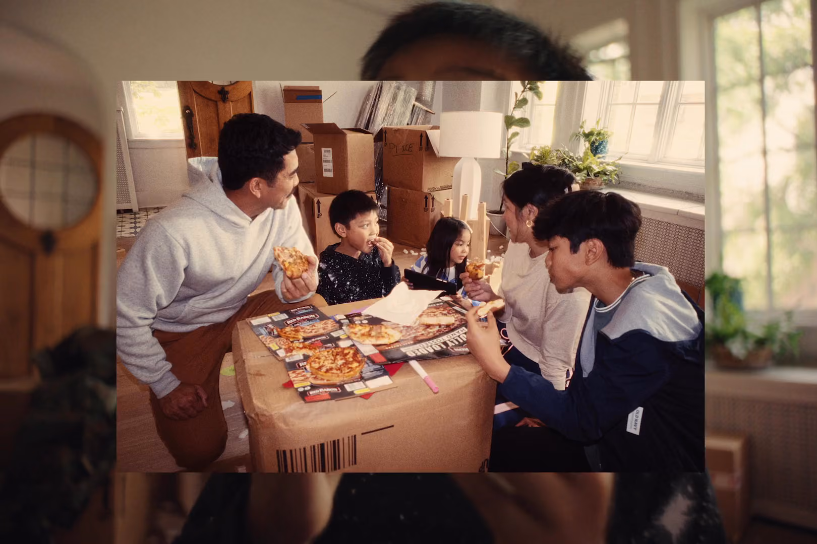 An image over another image. The image in the front shows a family of five eating pizza together. They are sat around a box, using it is a table.