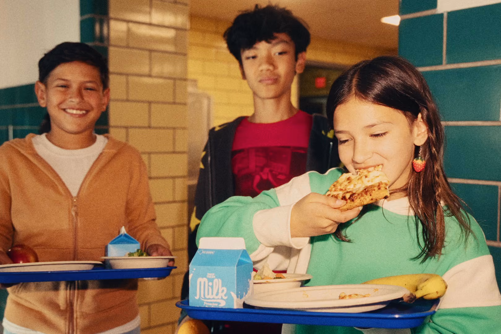 Two boys and a girl standing and holding trays of food. The girl is taking a bite of a pizza slice.
