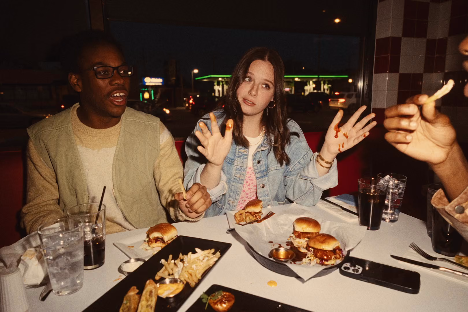 Two men and a woman eating burgers around a table in a restaurant. The woman is holding up her hands which have sauce from the food on them.