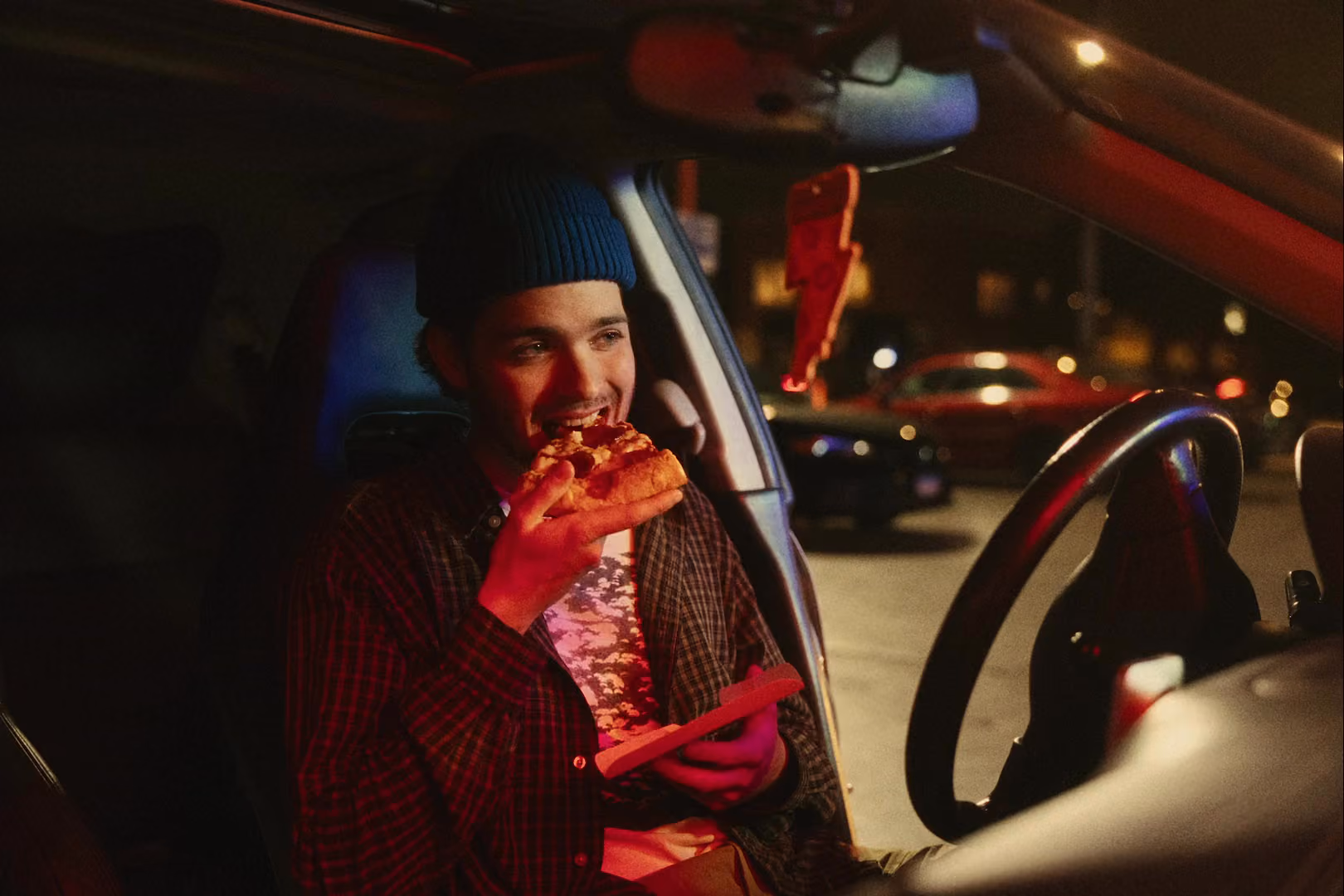 A man in a beanie eating pizza in a car.