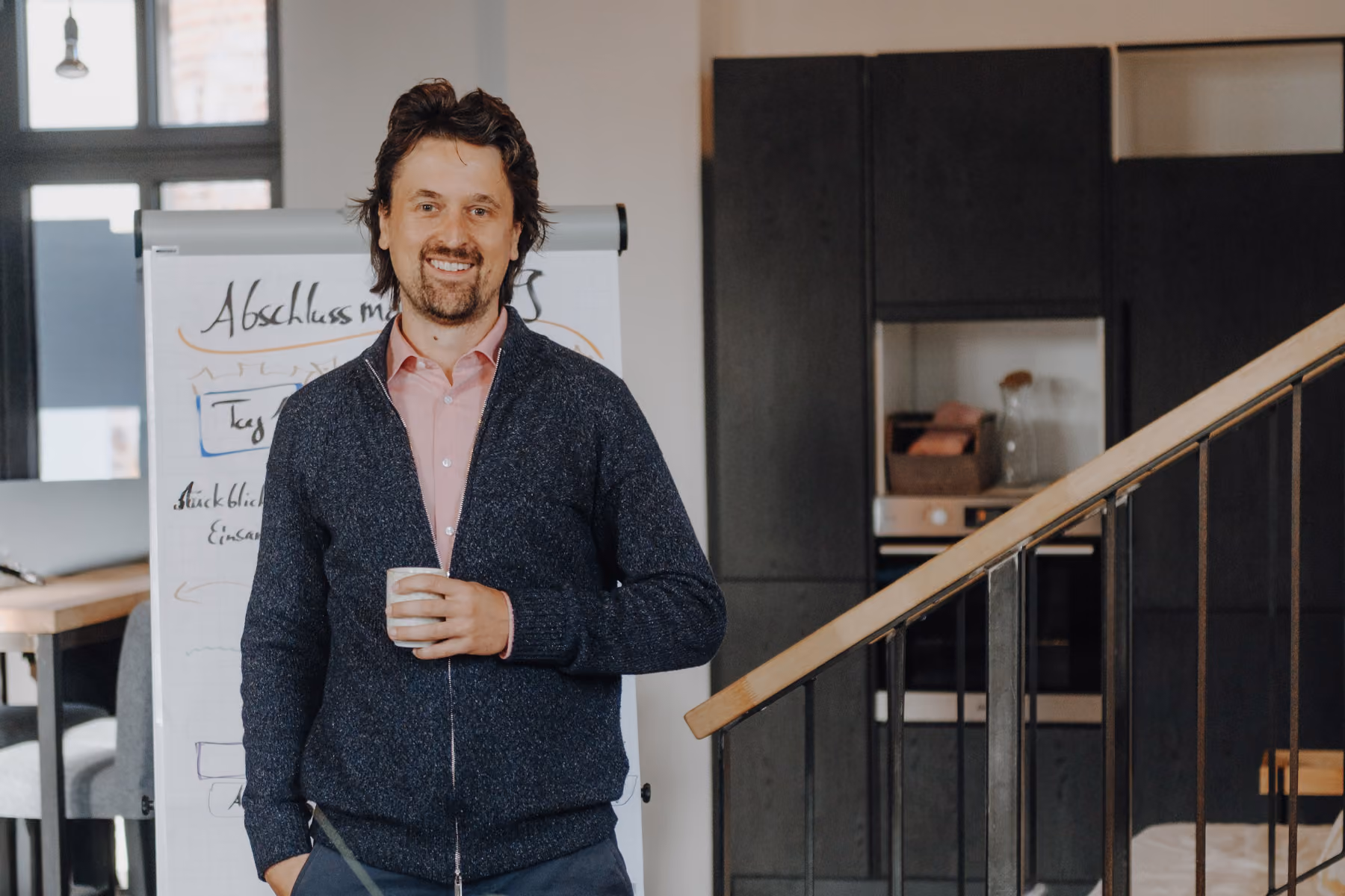 Dominik Frisch lächelt mit Kaffeetasse in der Hand vor einem Flipchart in einem modernen Büro.