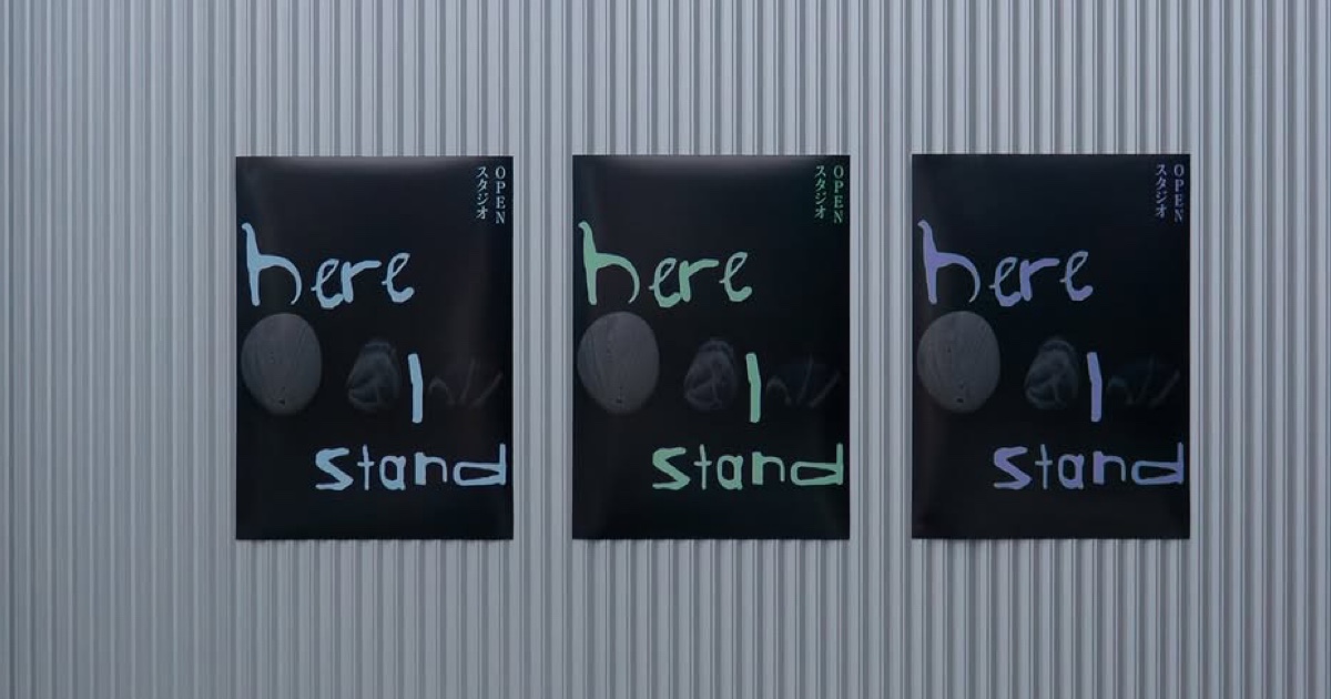 Three black posters on a corrugated metal wall displaying the phrase 'here I stand' in different pastel colors with small vertical text in Japanese and the word OPEN.