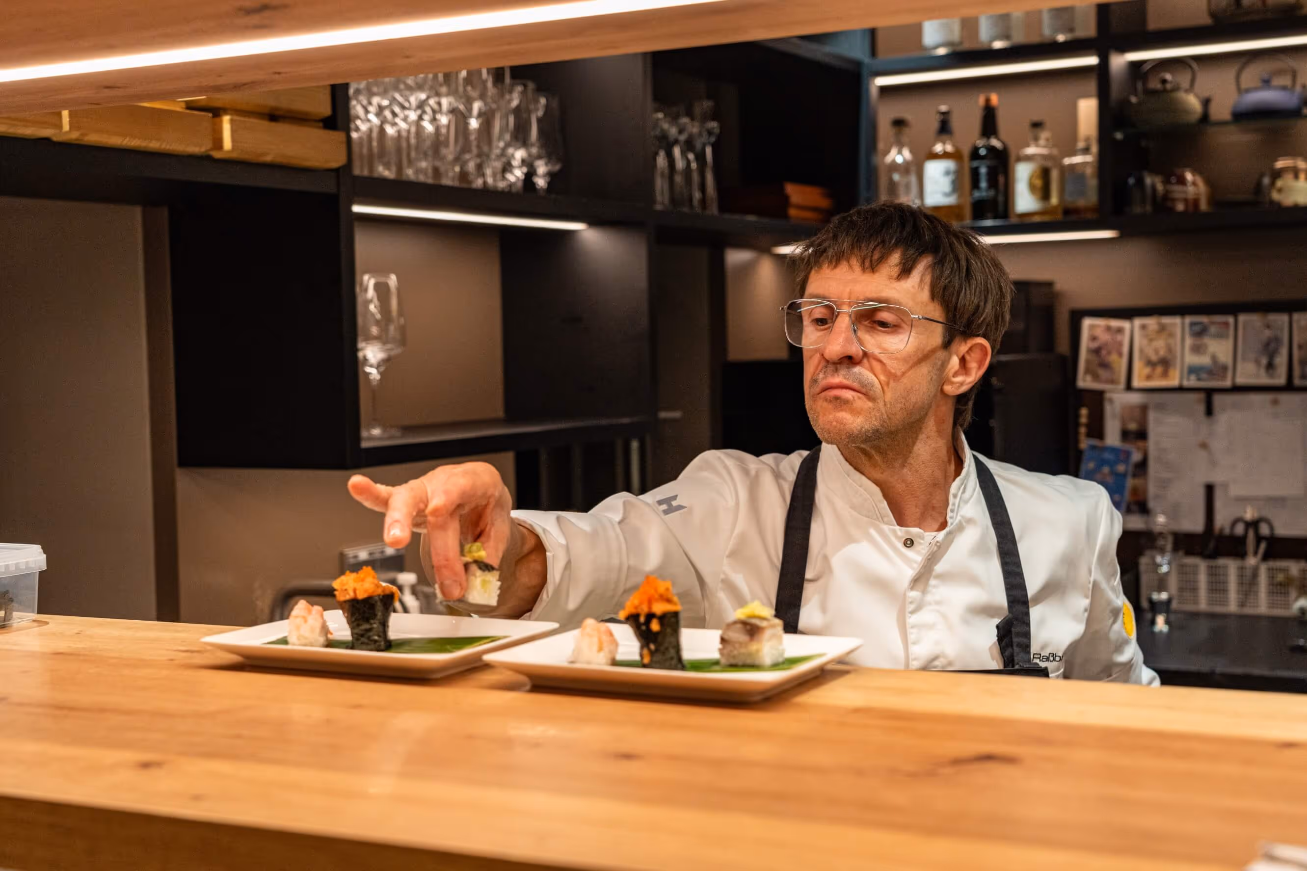 A sushi master wearing glasses and a white coat arranges pieces of sushi on plates in the restaurant kitchen at Nashi Dining in Erfurt.